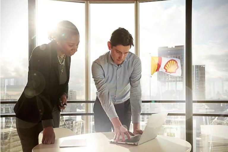 Two people looking at a laptop, flag with Shell logo can be seen out the window behind them